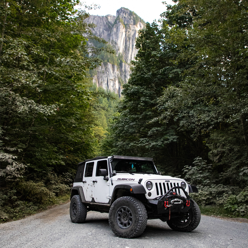 White Jeep surrounded by evergreens, mountains, on a gravel road.